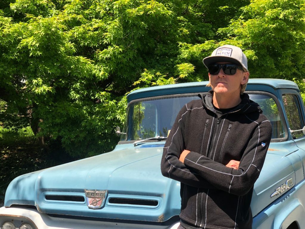 A man in a hoodie and a hat standing next to a blue truck thinking about life.