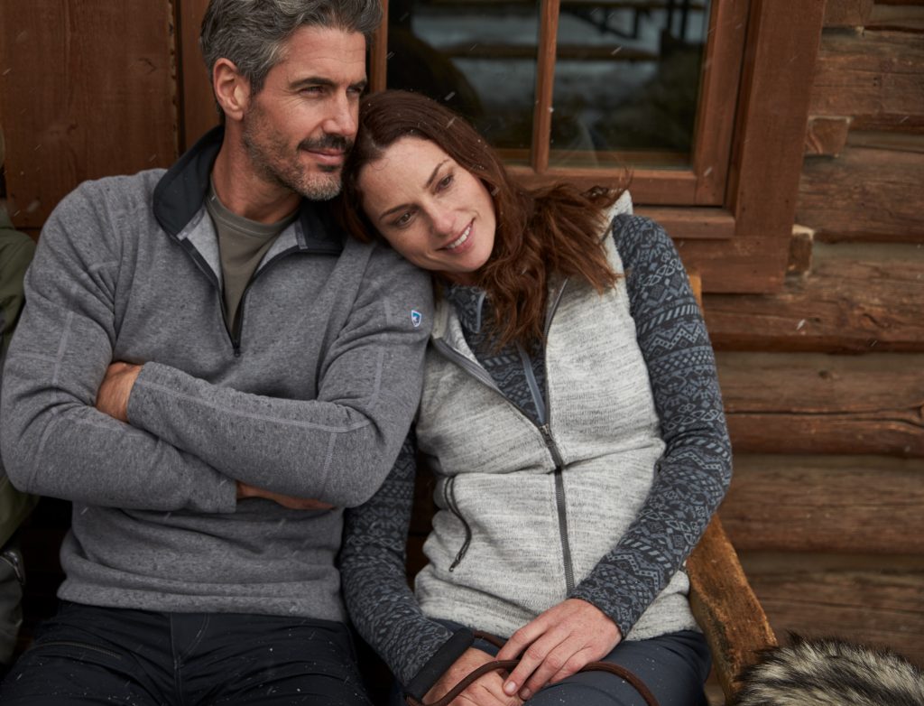 Woman leans on a man in front of a wooden cabin, dressed in KUHL winter clothing.