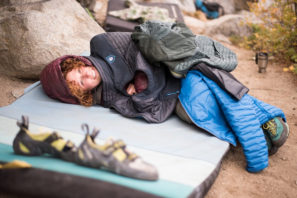 A smiling woman laying on a mat covered with multiple KUHL women's jackets.