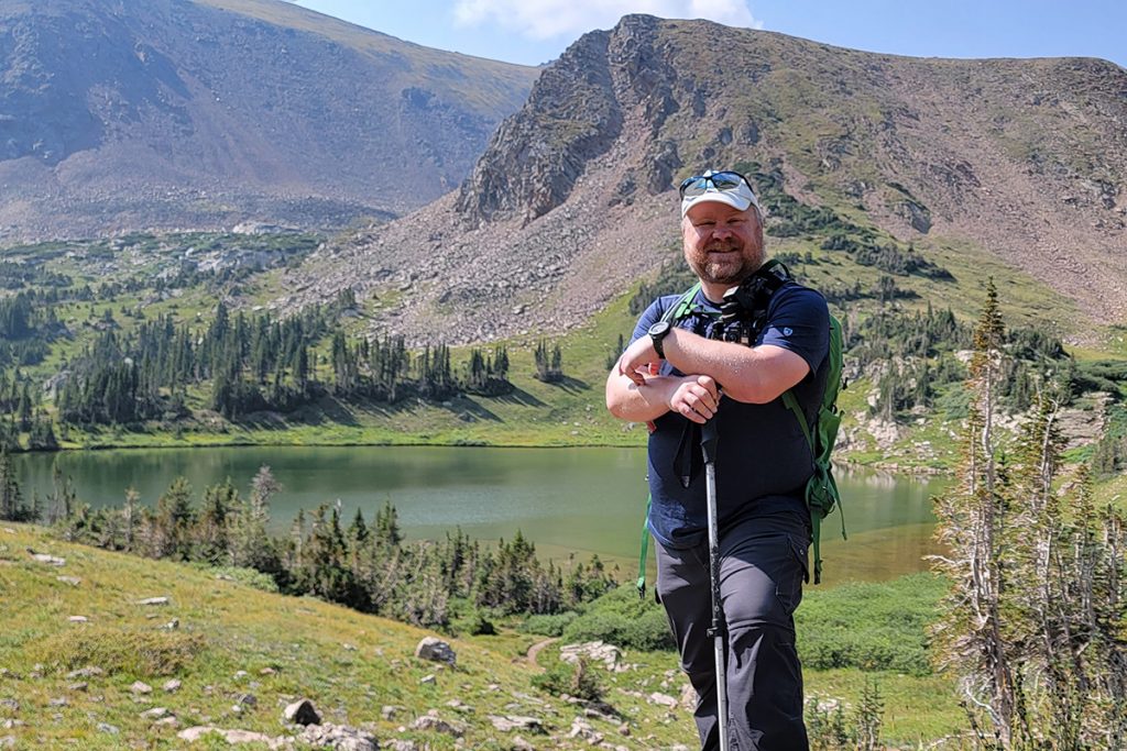 man in KÜHL shirt standing in front of the lake during daytime